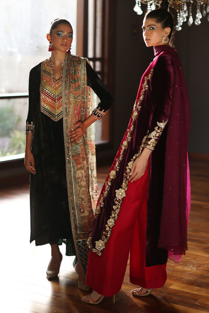Two women in traditional outfits standing indoors with a window in the background.