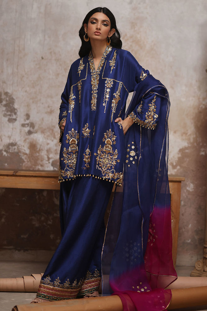 Woman wearing a traditional blue embroidered outfit with a pink dupatta against a textured wall.