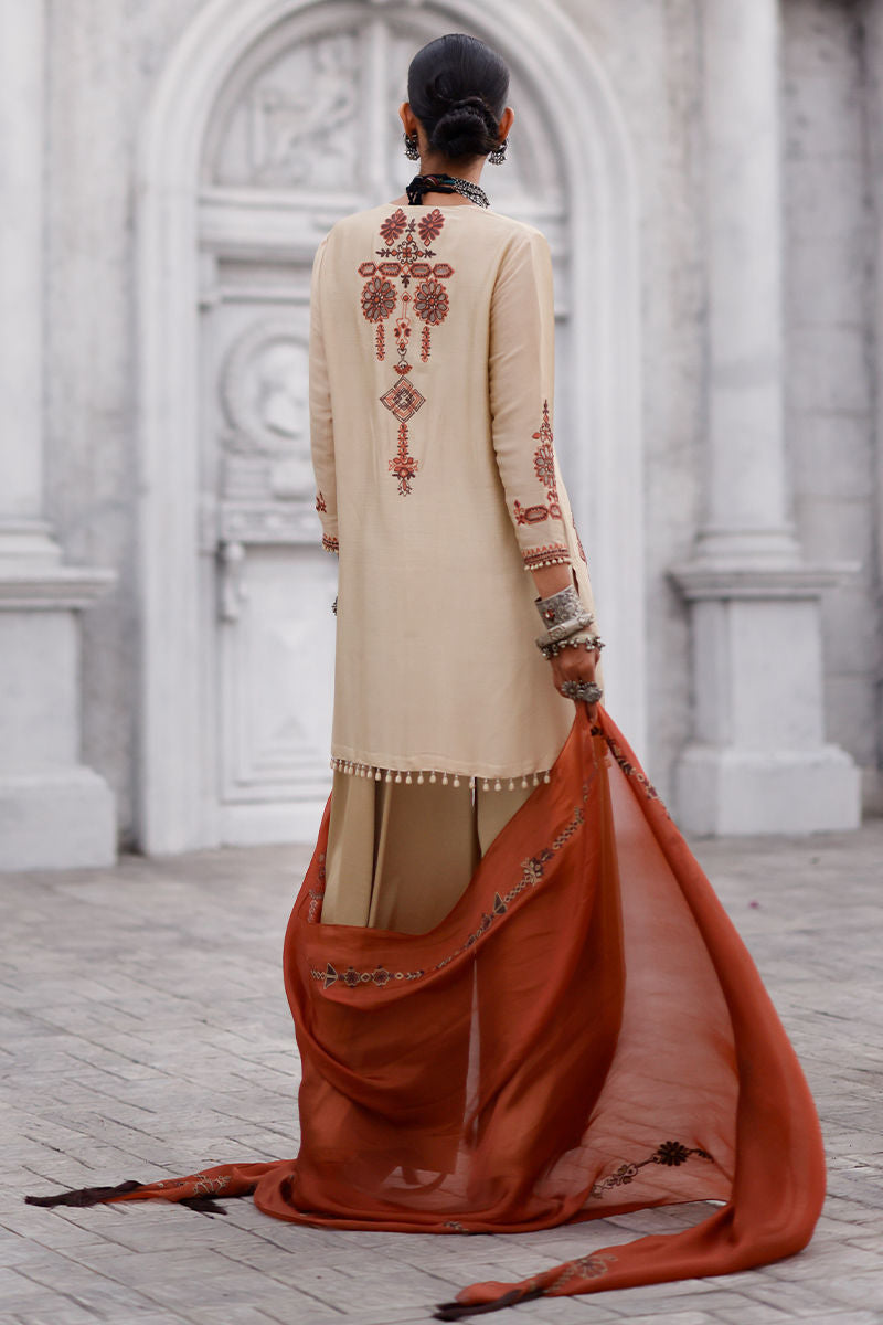 Person wearing a beige and orange traditional outfit with intricate patterns in an elegant setting.