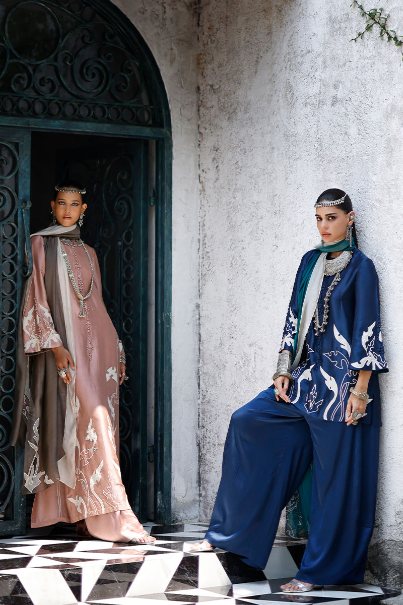 Two women in traditional outfits standing against a white wall with decorative elements.