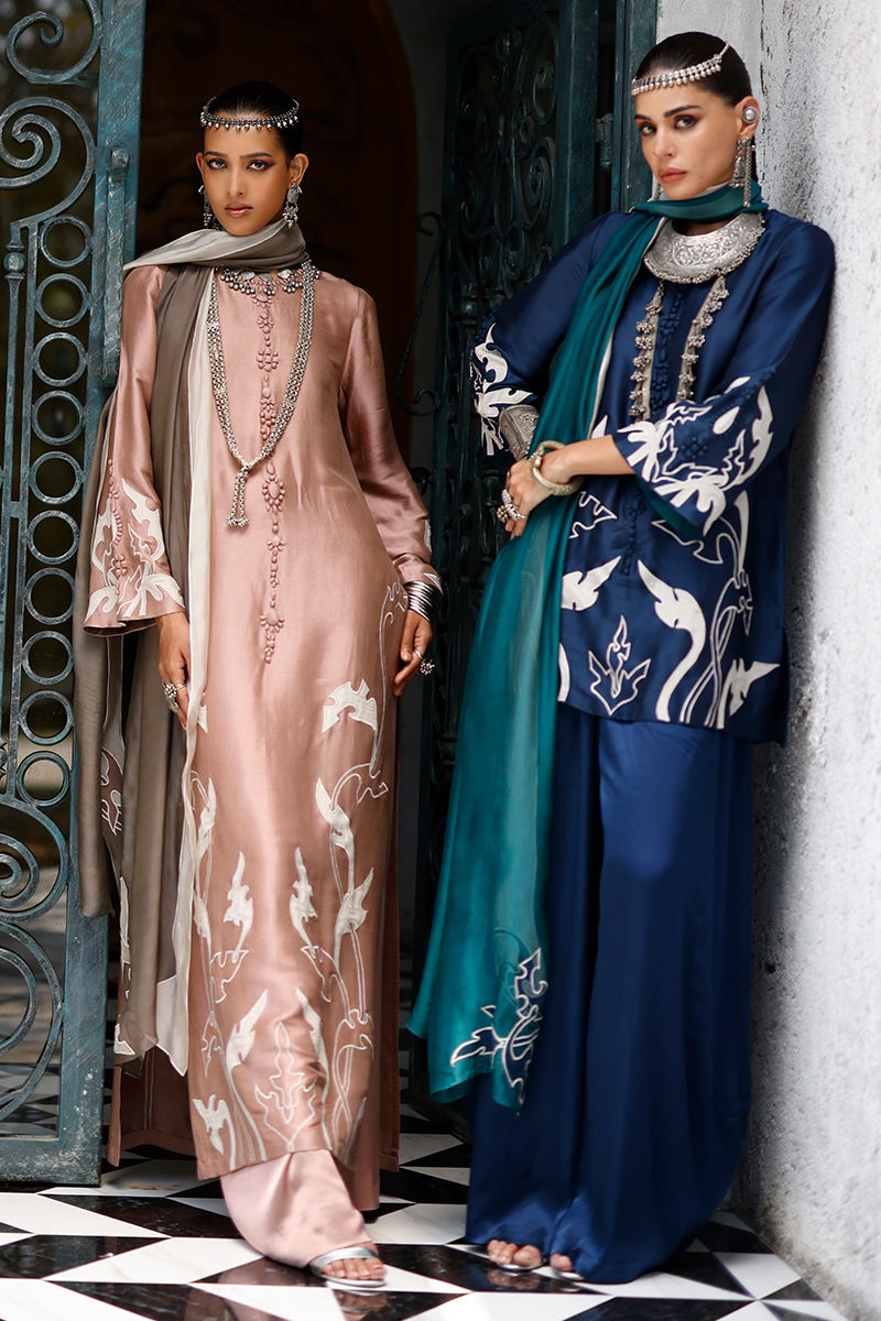 Two women in traditional embroidered dresses standing against a decorative wall.