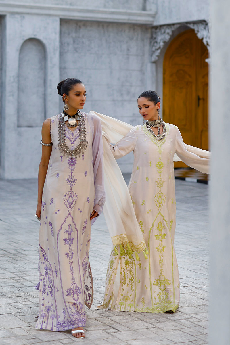 Two women in elegant dresses with intricate designs standing in a decorative outdoor setting.