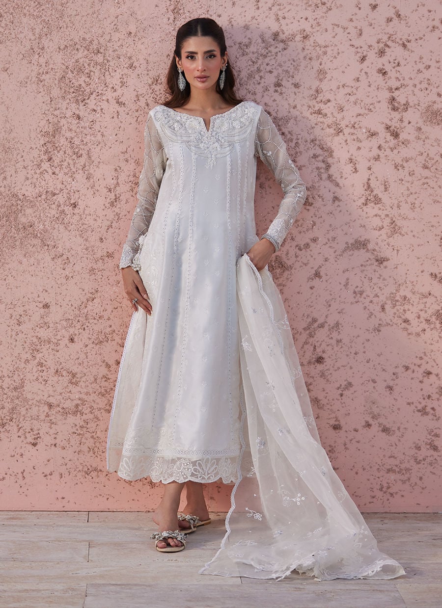 Woman wearing a white embroidered dress against a pink textured wall.