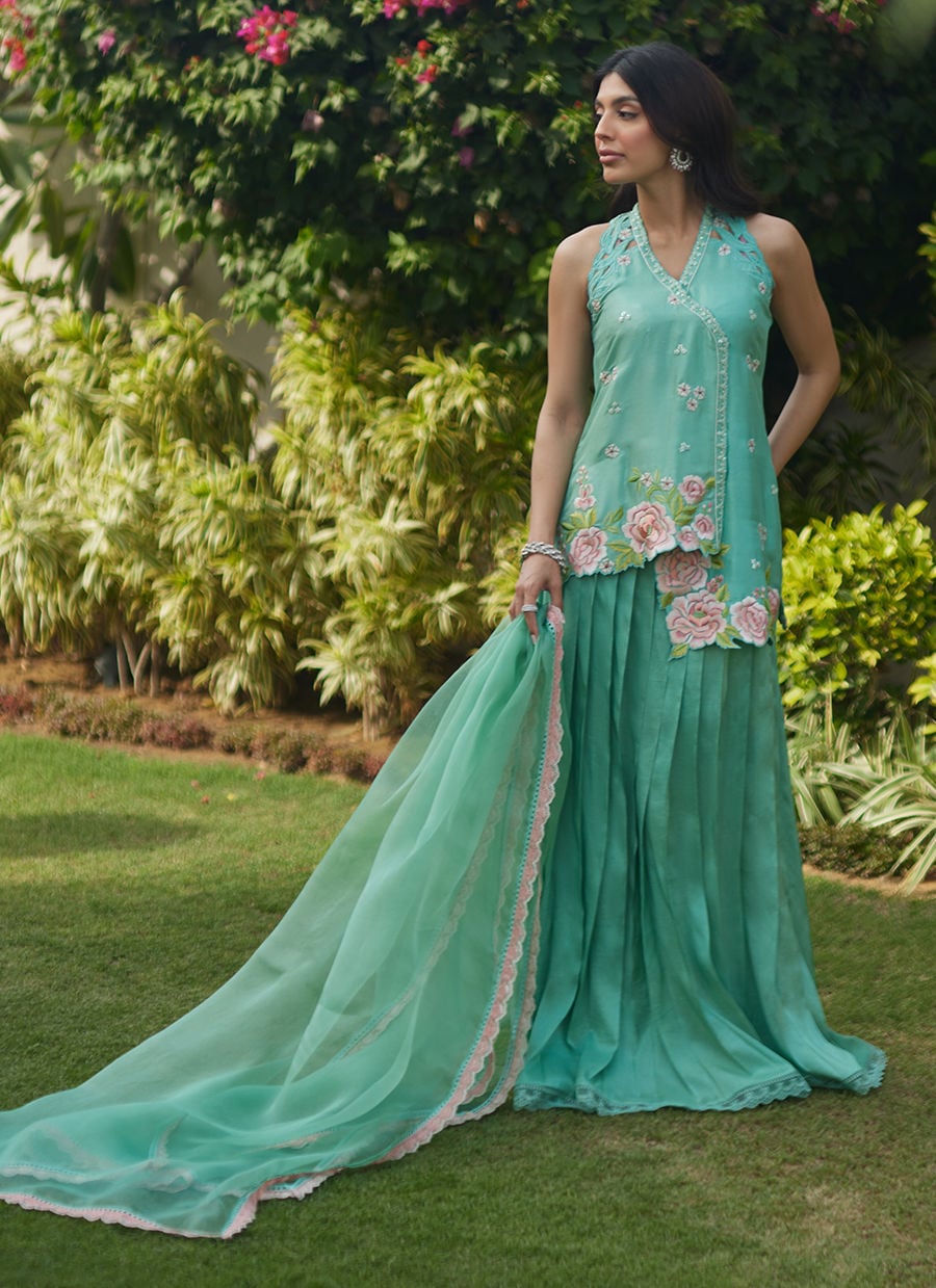Woman in a teal saree with floral patterns standing outdoors among greenery