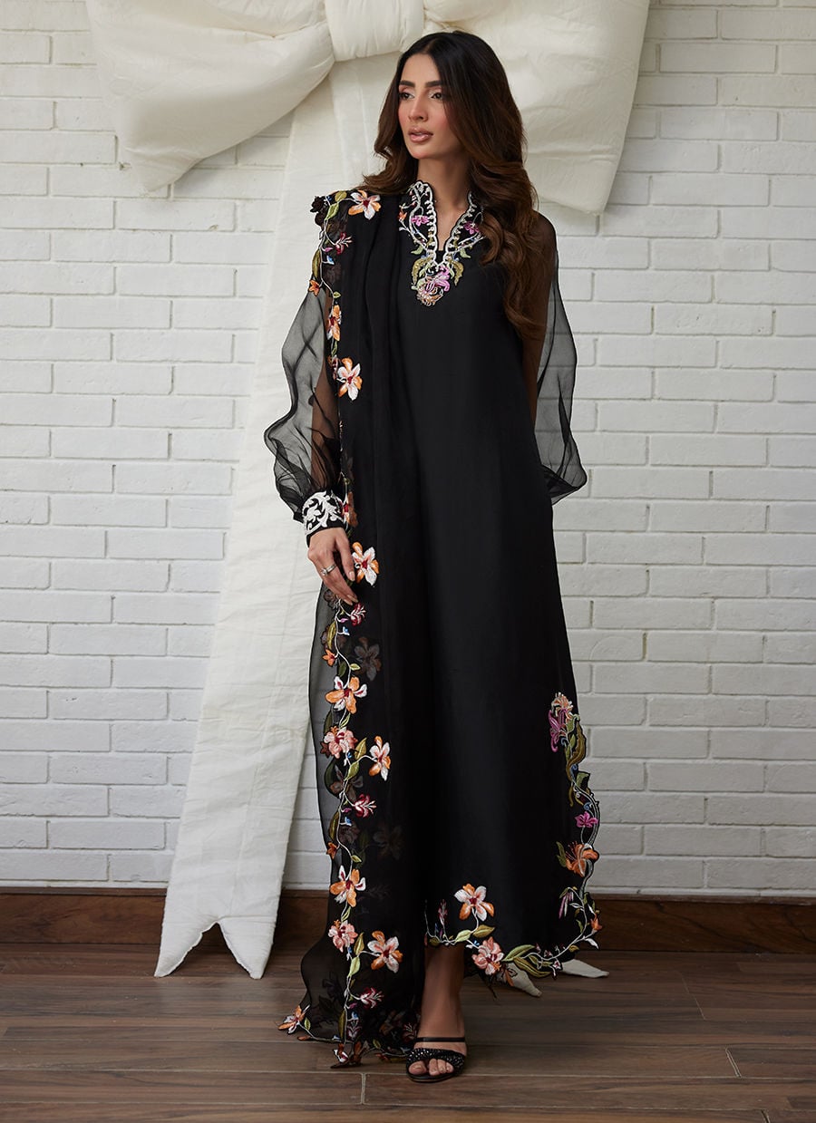 Woman wearing a black dress with floral embroidery against a white brick wall.