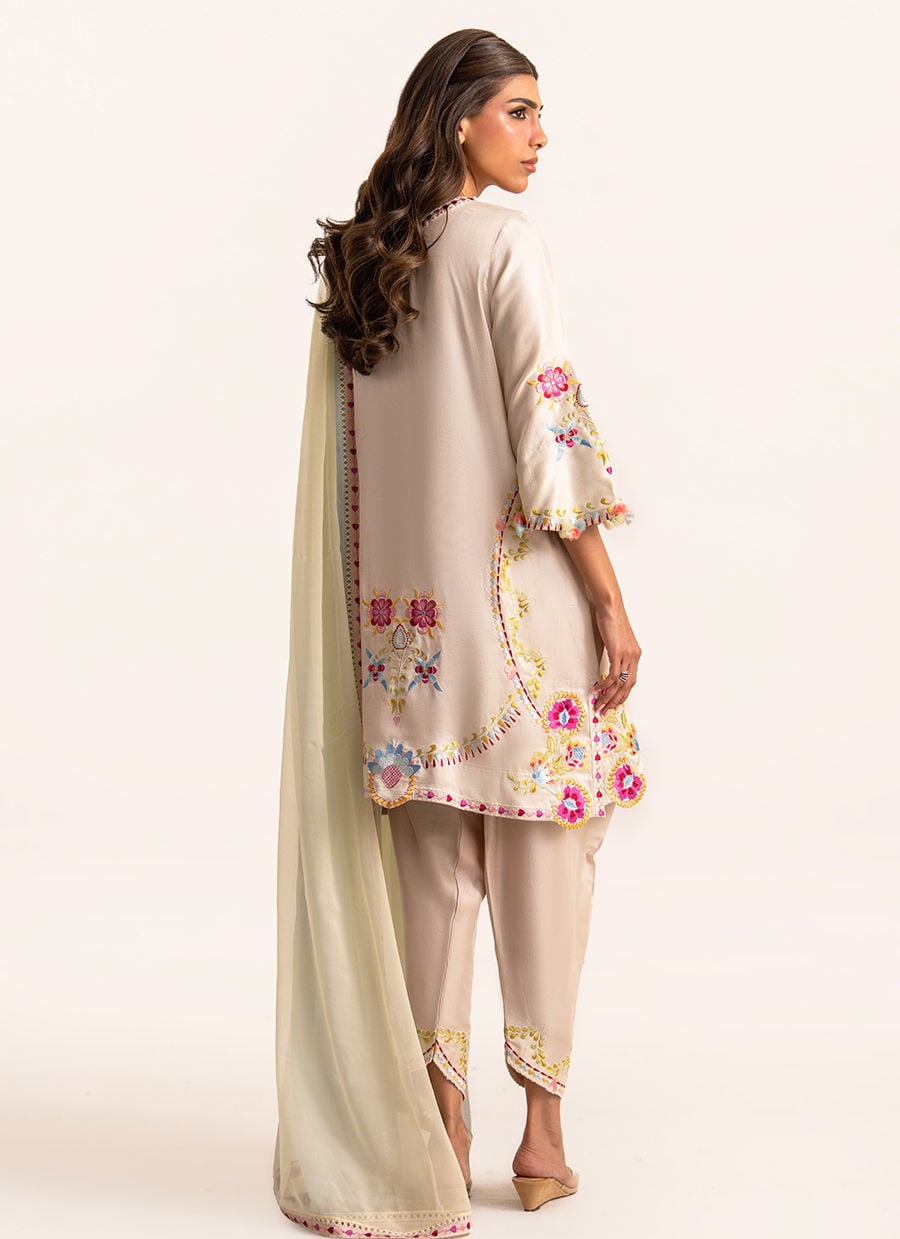 A woman modeling a traditional three-piece salwar kameez suit in light beige, featuring a long kurta with colorful floral embroidery on the sleeves and hem, matching trousers, and a draped dupatta, shown from a side-back angle against a plain cream background.