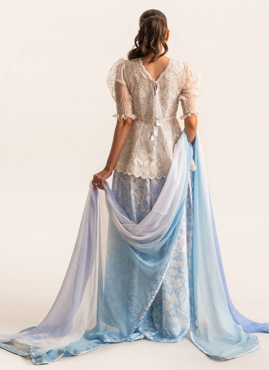 A woman shown from the back wearing a traditional outfit with a cream lace top and a flowing light blue patterned skirt, holding a sheer blue and white dupatta against a plain light background.