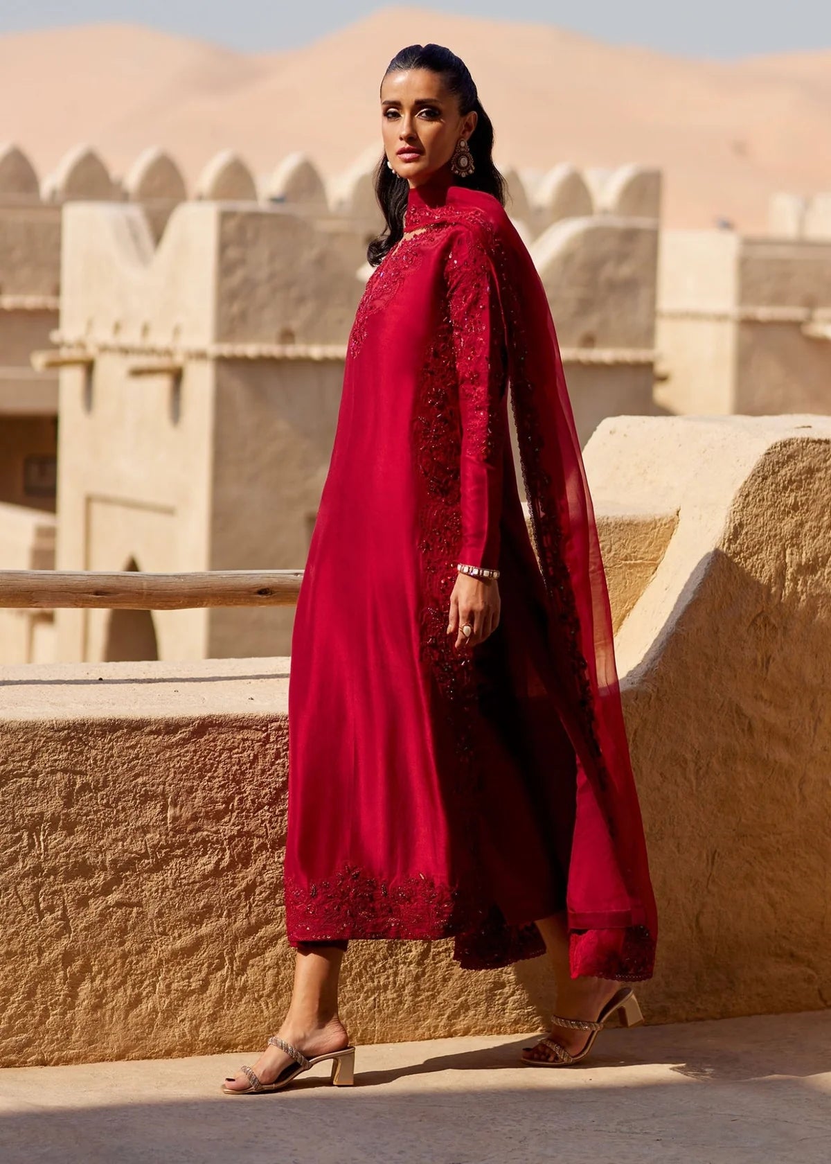 Woman in a red traditional outfit standing in front of a sandy architectural structure.