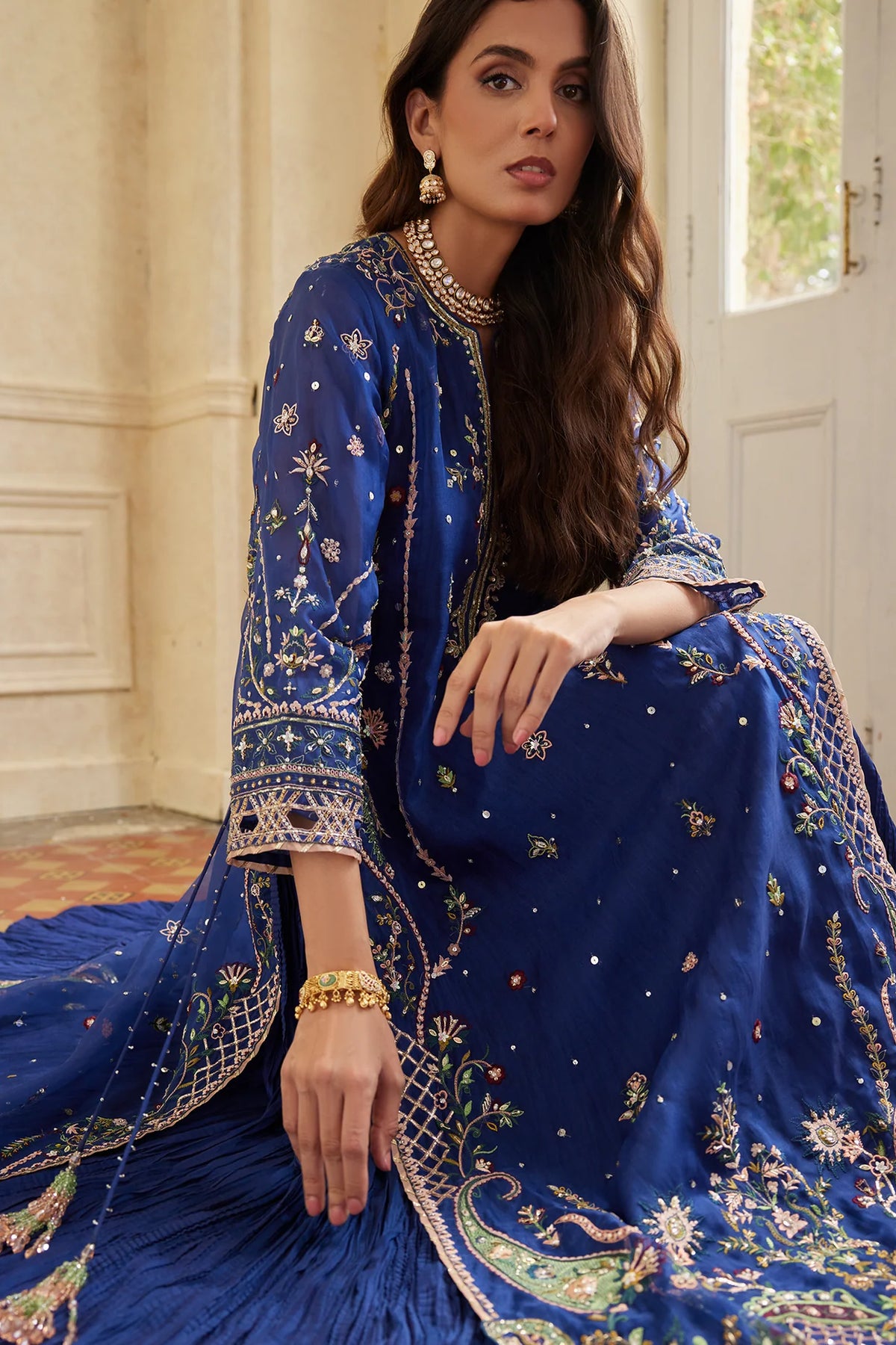 Woman in a blue embroidered traditional outfit sitting indoors.