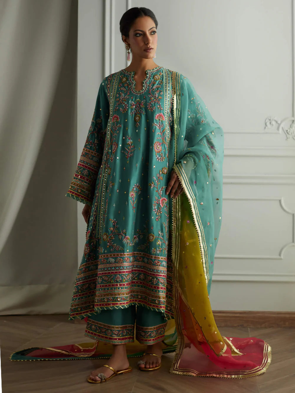 Woman wearing a traditional outfit with a teal embroidered dress and dupatta in a room.