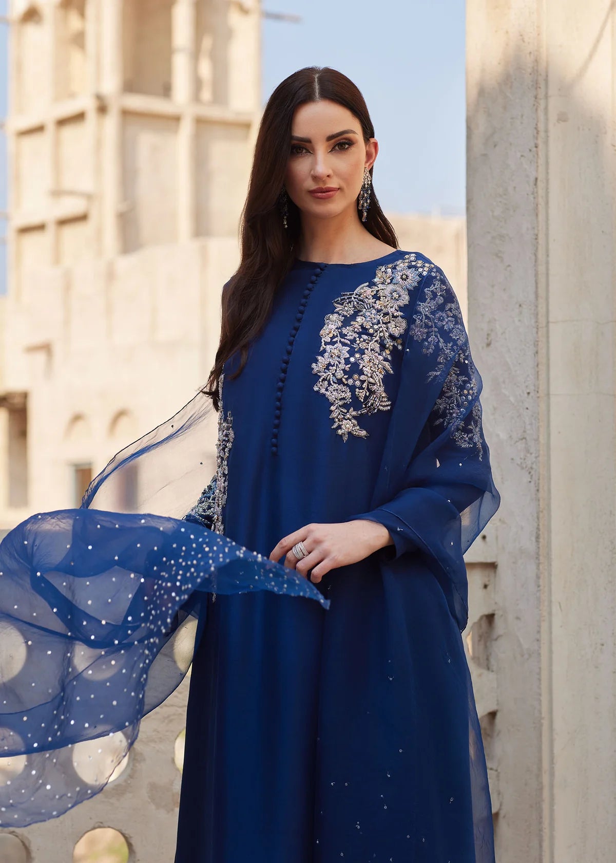Woman wearing a blue dress with intricate white embroidery against a blurred architectural background
