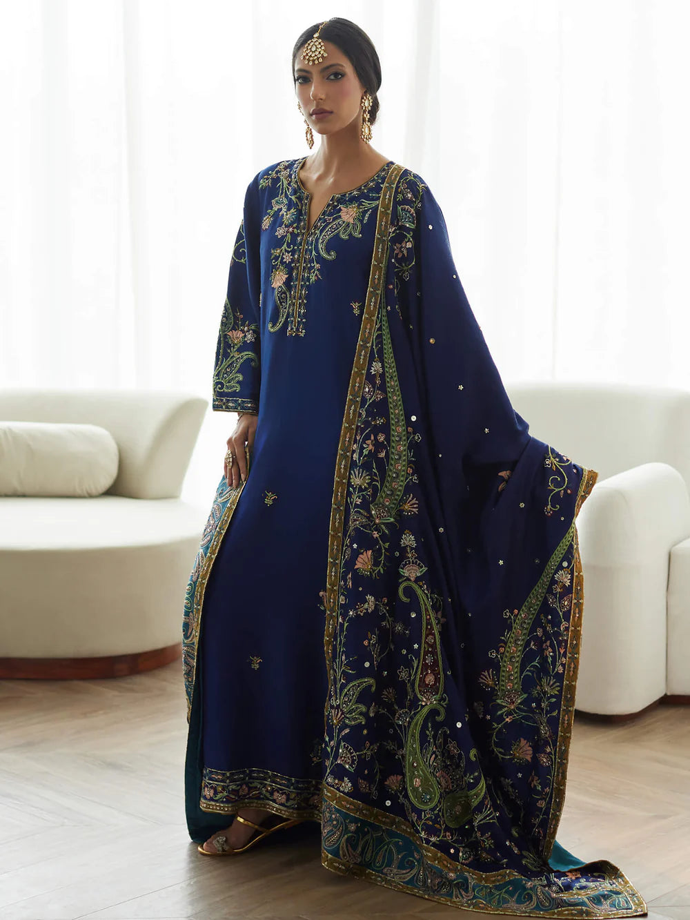 Woman wearing a blue embroidered traditional outfit in a room with white sofas.