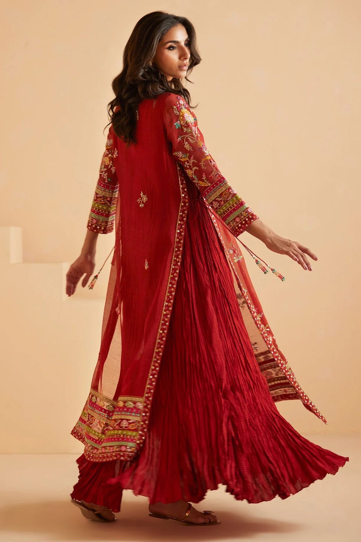A woman modeling a traditional red Indian outfit with gold and green embroidery, featuring a long kurta, pleated skirt, and matching dupatta against a warm beige background.