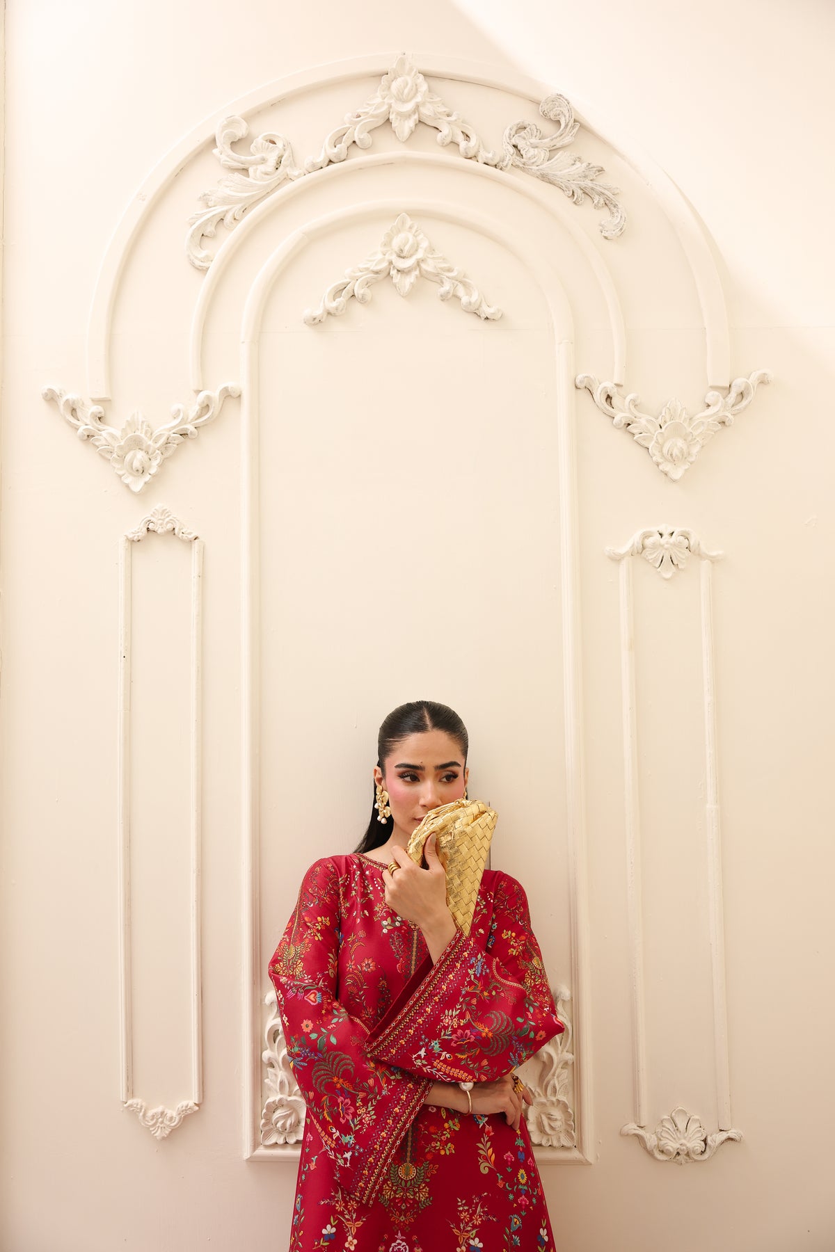 Woman in a red traditional outfit holding a gold item against a decorative white wall.