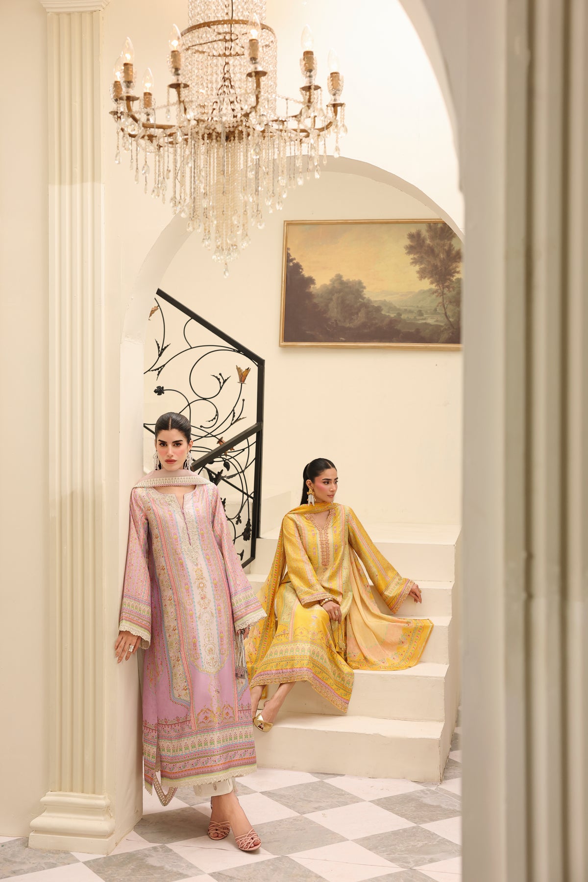 Two women in traditional outfits standing in a elegant interior setting with a chandelier.