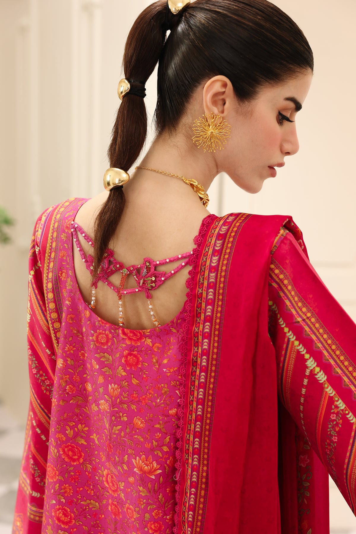 Woman wearing a pink and red traditional outfit with gold jewelry against a neutral background
