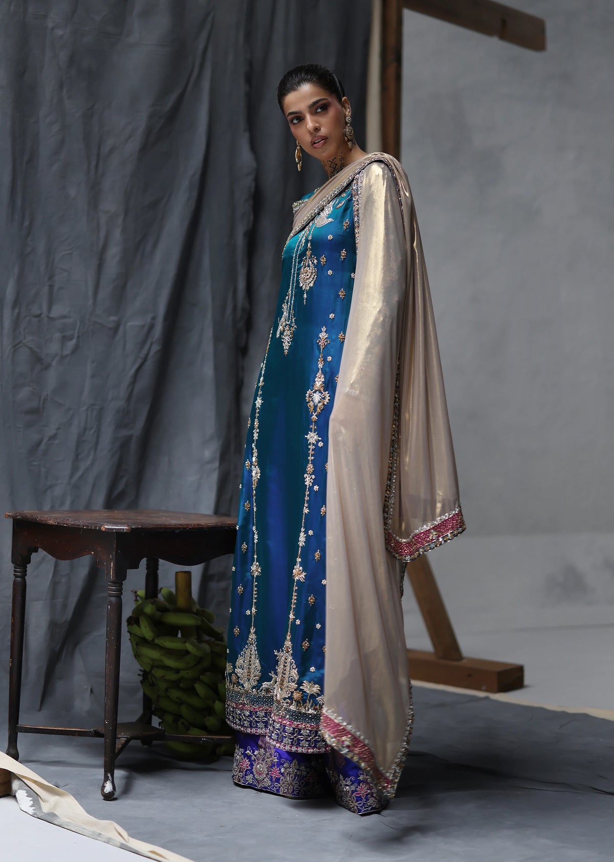 A woman models a traditional Indian outfit consisting of a teal blue kurta heavily embroidered with gold and silver threadwork, paired with a beige dupatta and a purple border skirt, set against a gray studio backdrop.