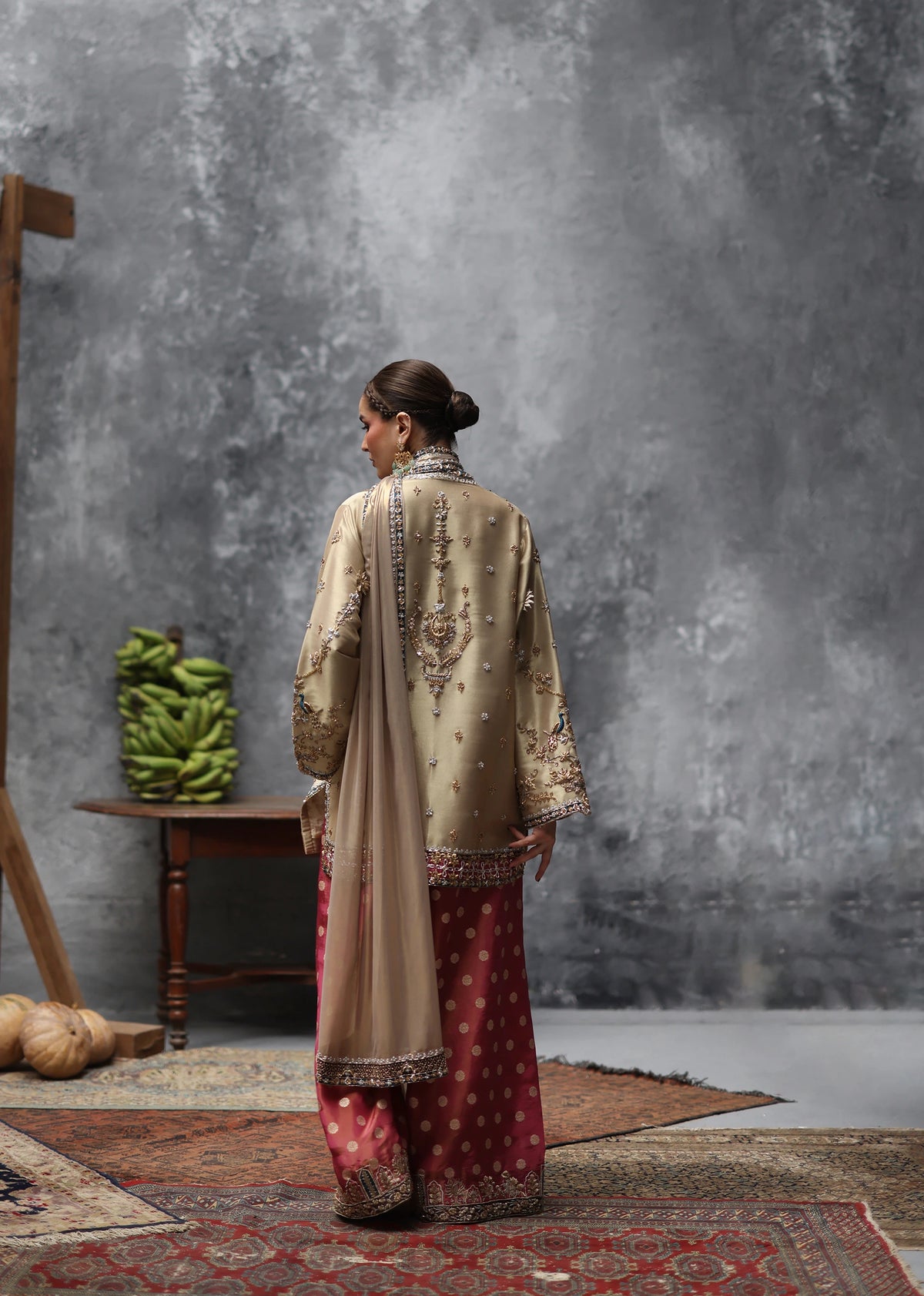 A woman stands with her back to the camera wearing a traditional beige embroidered kurta and a red patterned skirt, posed in a rustic studio setting with a textured gray wall, wooden table, and patterned rugs.