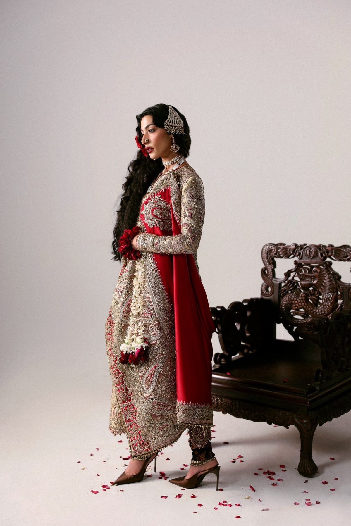 Woman in traditional outfit with red and white patterns standing next to an ornate chair on a light background.
