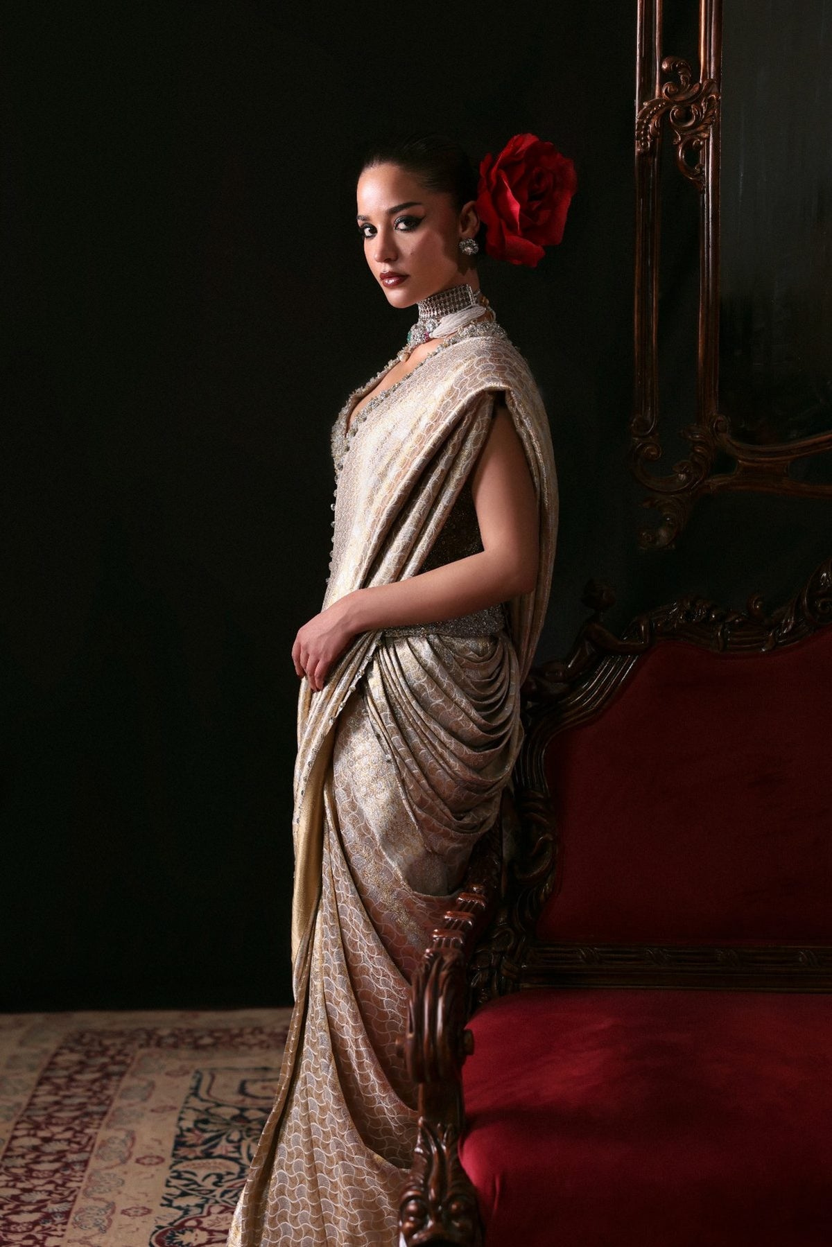 Woman in a traditional saree with a red flower in her hair, standing against a dark background.
