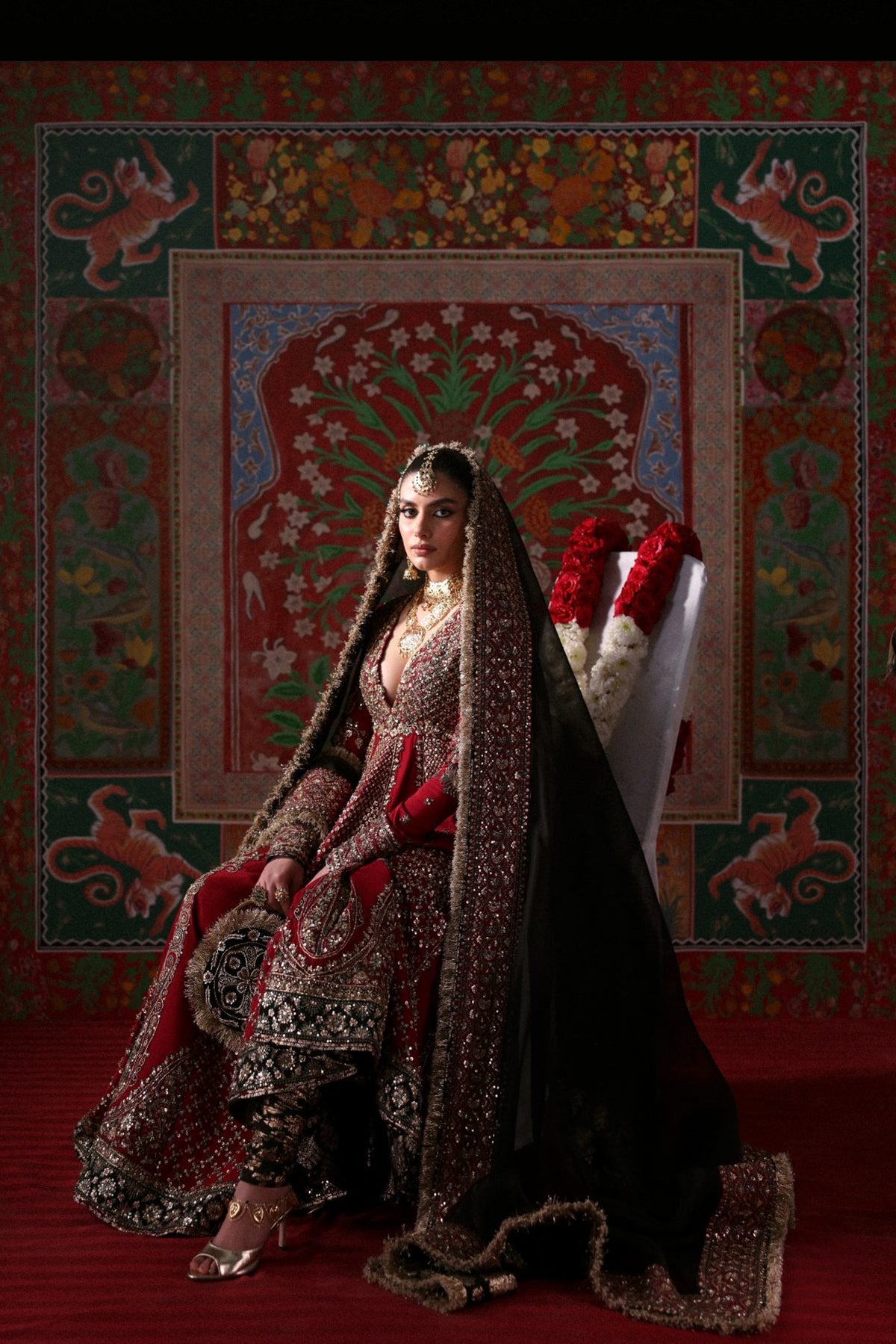 Woman in traditional attire sitting on a decorative chair with a colorful patterned wall behind her.
