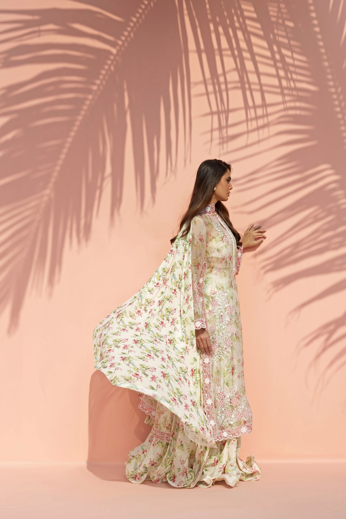 Woman in a floral dress standing against a pink wall with palm leaf shadows