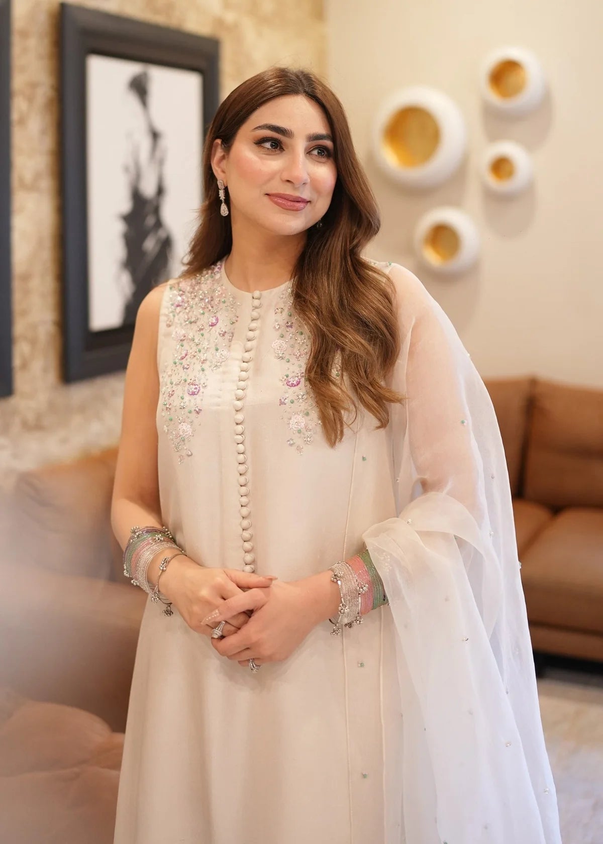 Woman in a white embroidered outfit standing in a decorated room with a couch and wall art.