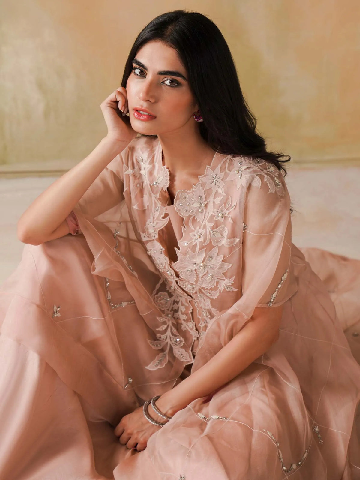 Woman in a peach-colored traditional outfit with floral embroidery sitting on a couch.
