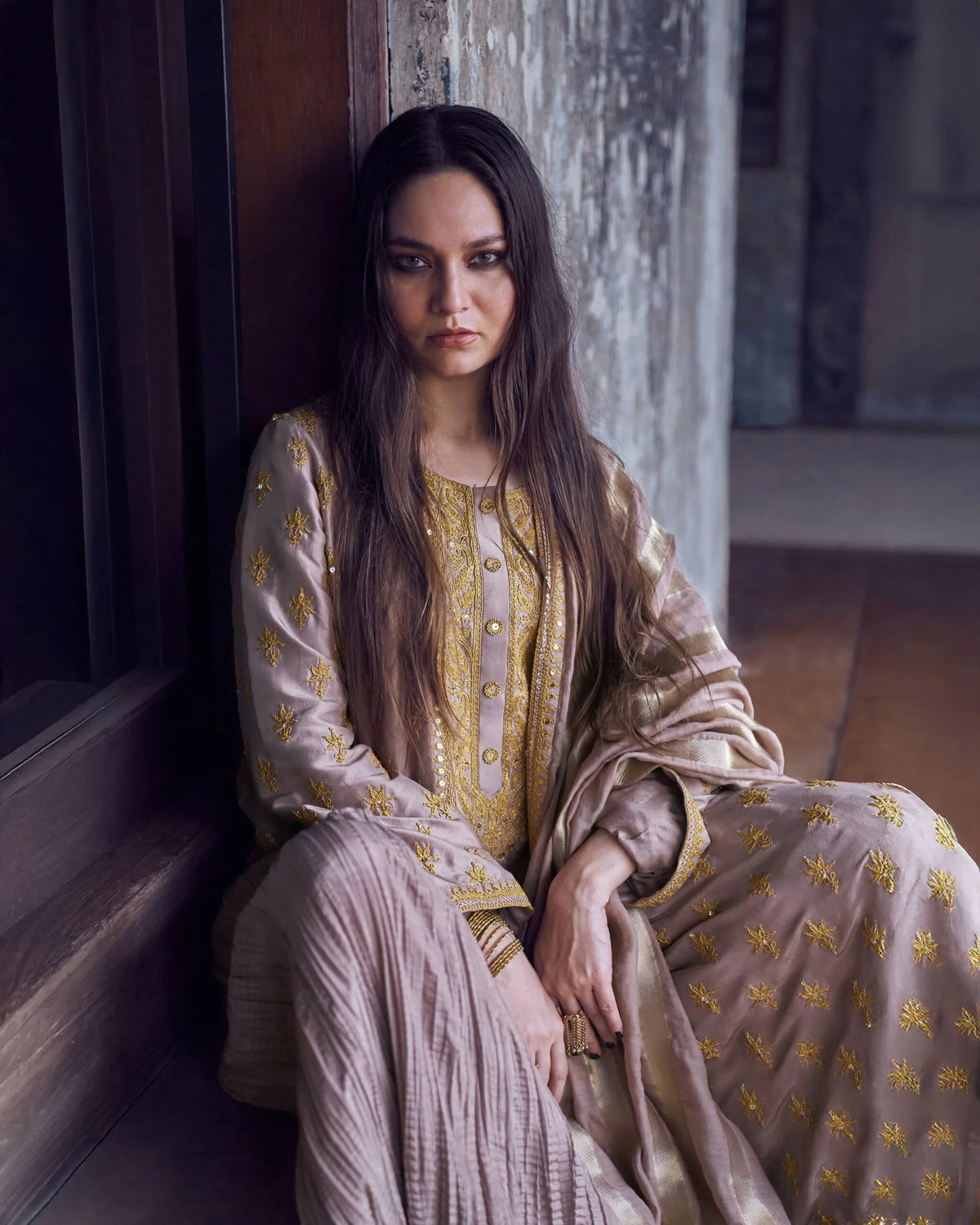 Woman in an embroidered traditional outfit sitting against a textured wall.