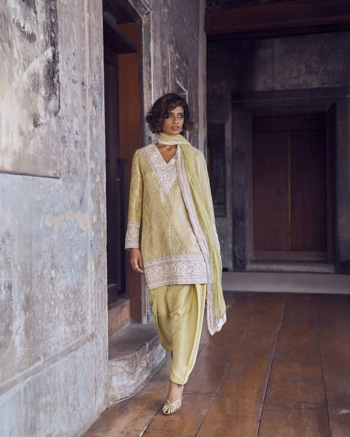 Woman in a light green traditional outfit standing in a rustic interior setting.