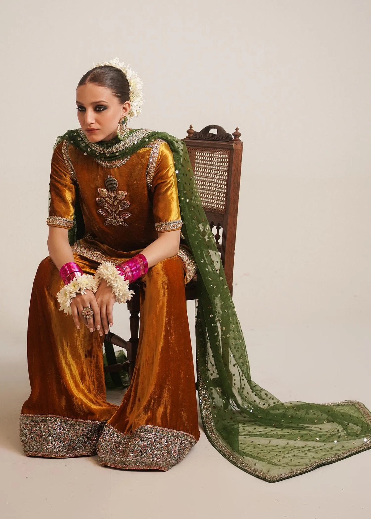Woman in traditional outfit with green dupatta sitting on a chair against a plain background