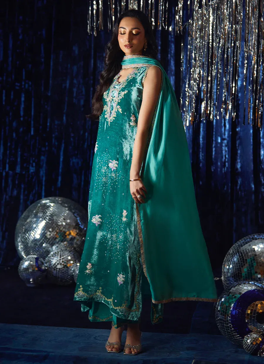 Woman in a teal embroidered outfit standing against a dark background with decorative elements.