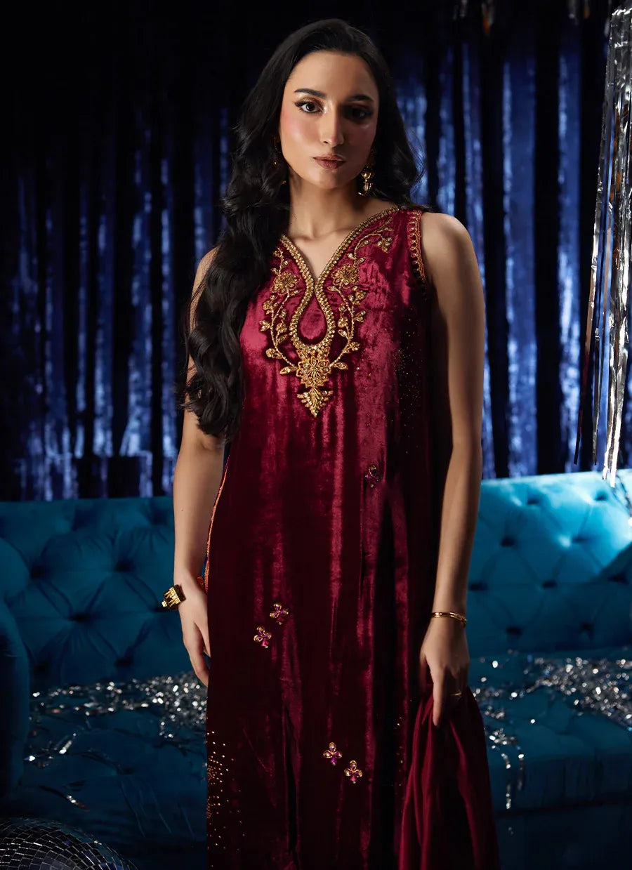 Woman in a burgundy dress with gold embroidery standing in front of a blue couch with decorative elements.
