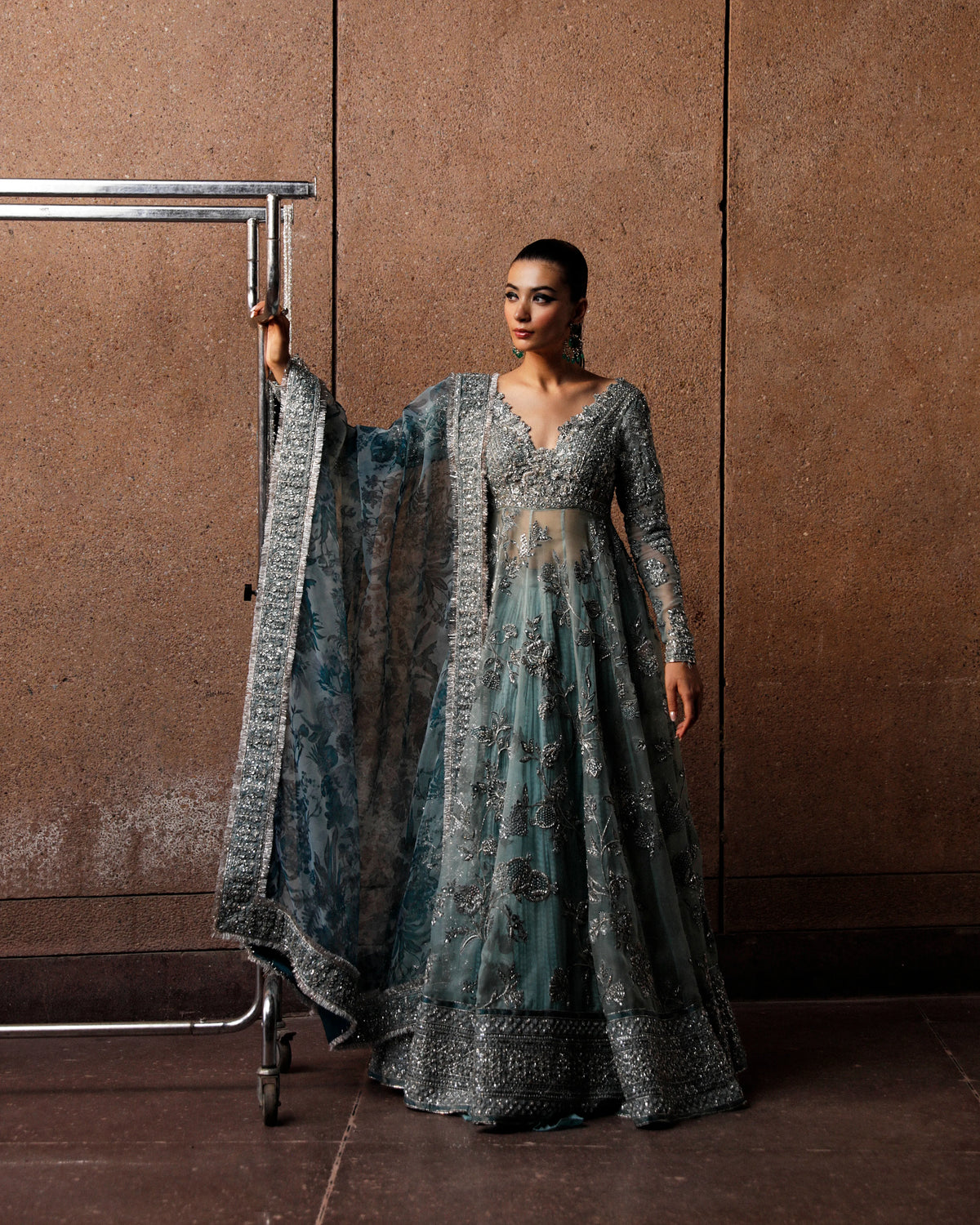 Woman in a long, elegant dress with intricate patterns against a brown wall.