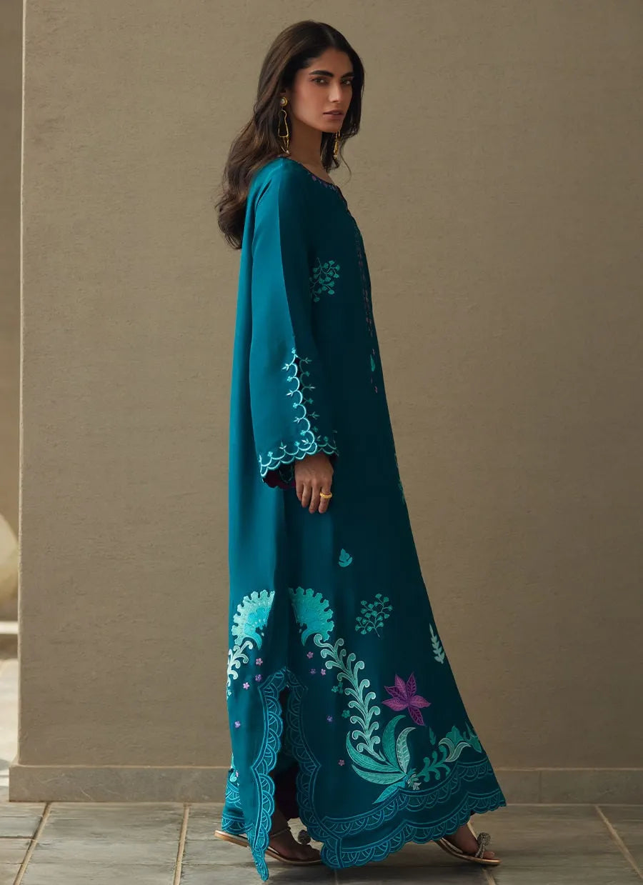 Woman wearing a teal embroidered dress against a beige wall.