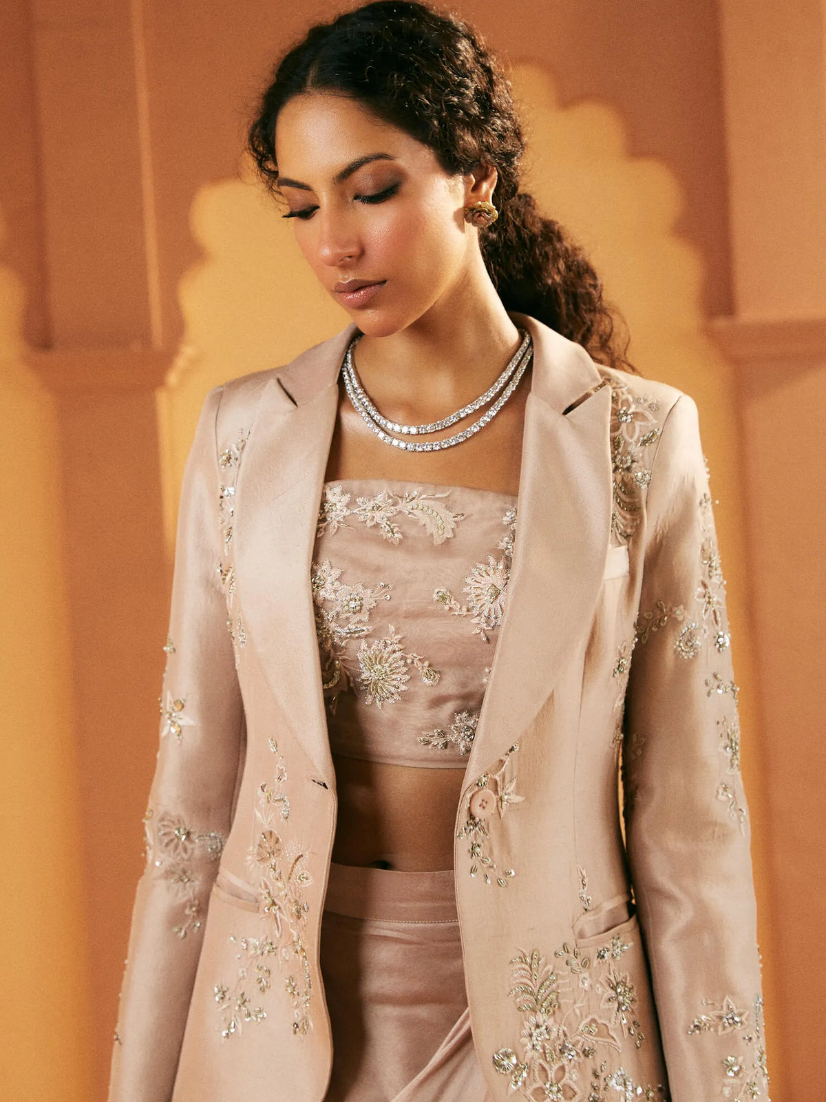 Woman wearing a beige embroidered outfit with a decorative necklace against an orange wall.