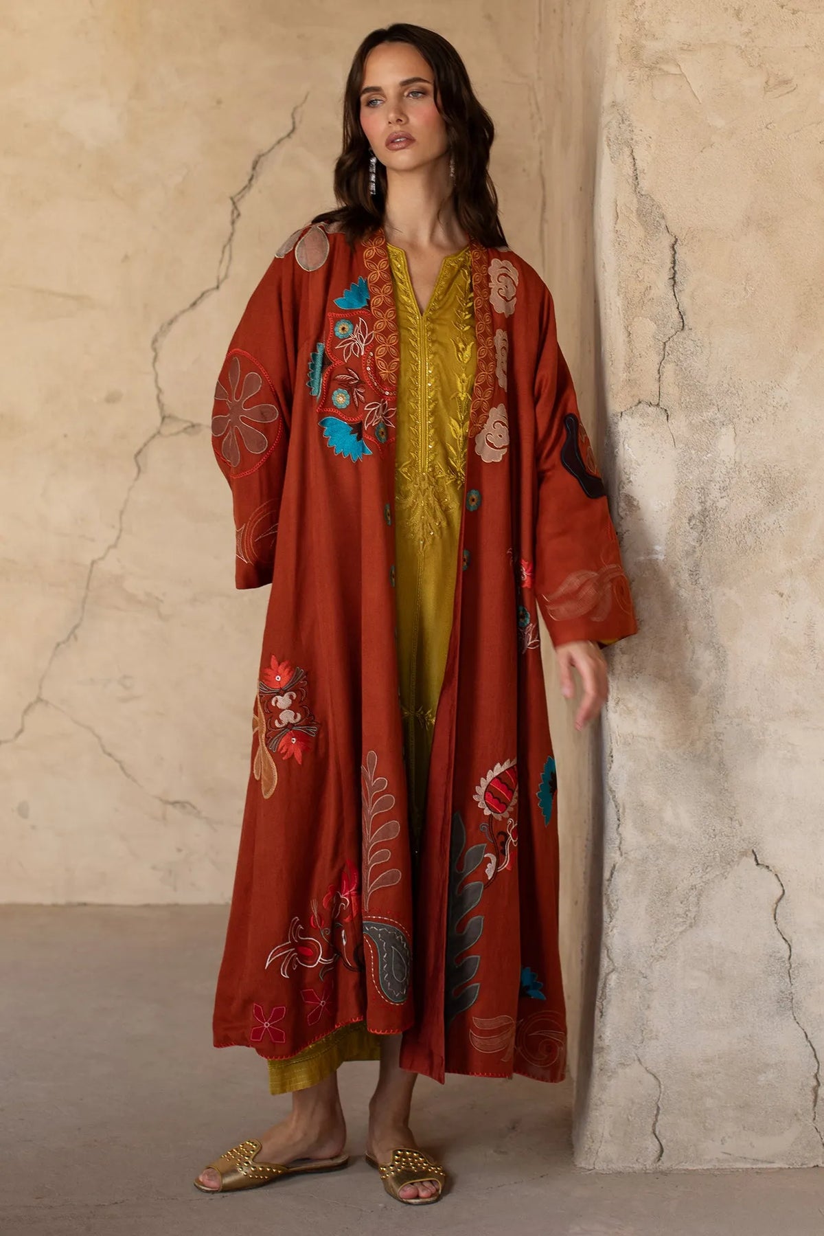 Woman wearing a red embroidered coat over a yellow dress against a textured wall.