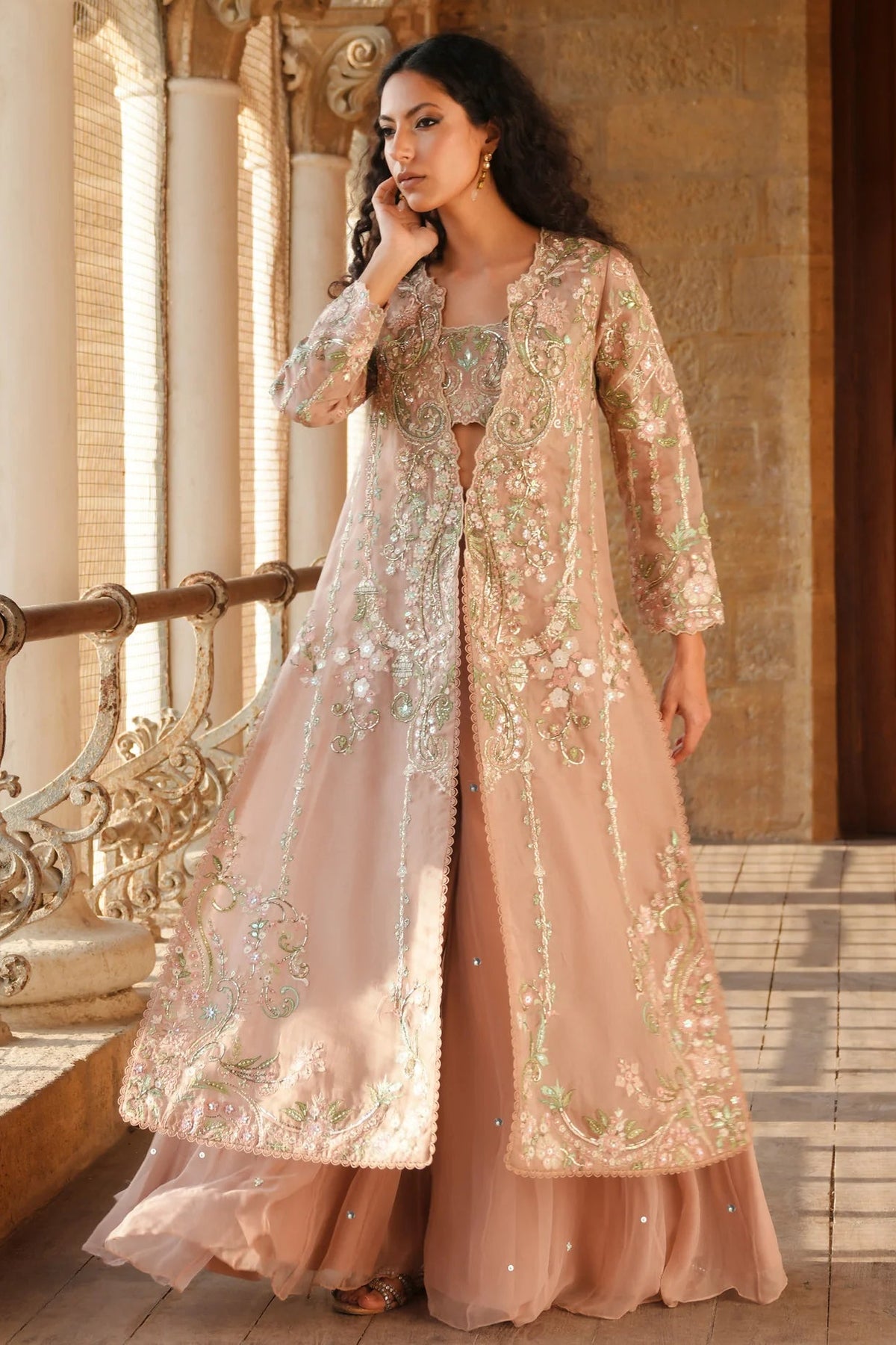 A woman models a traditional Indian outfit consisting of a peach-pink anarkali kurta with extensive silver and gold embroidery, paired with matching flared pants and a sheer matching jacket, set against an ornate stone corridor.