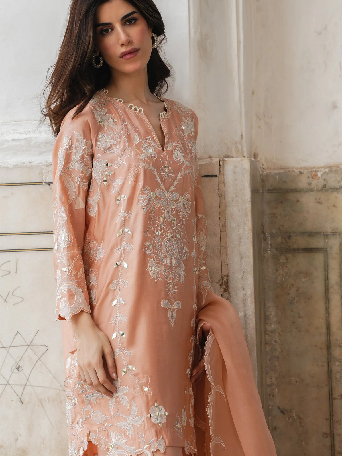 Woman wearing a peach embroidered dress against a textured wall.