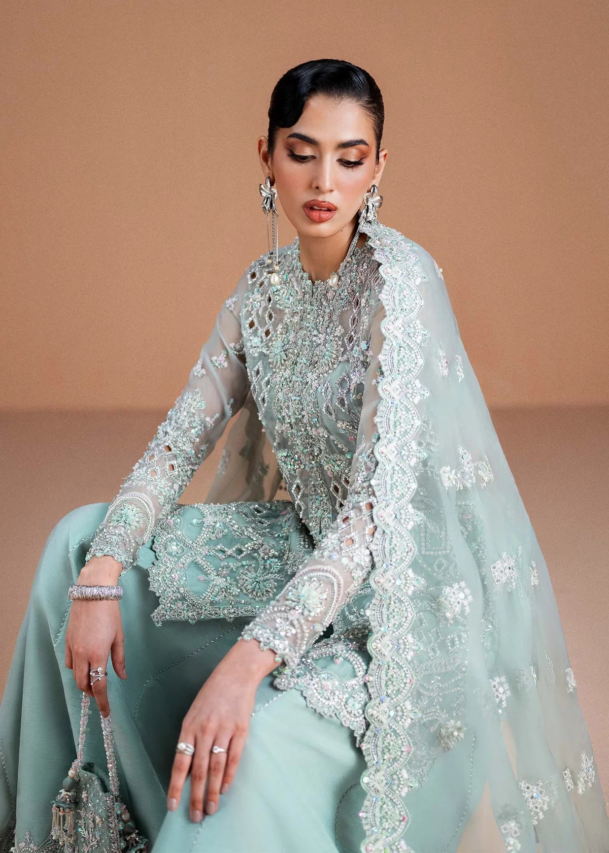 A woman seated and wearing an ornate mint green traditional outfit with heavy silver embroidery, sheer sleeves, a matching embroidered dupatta, and matching jewelry against a warm beige studio background.