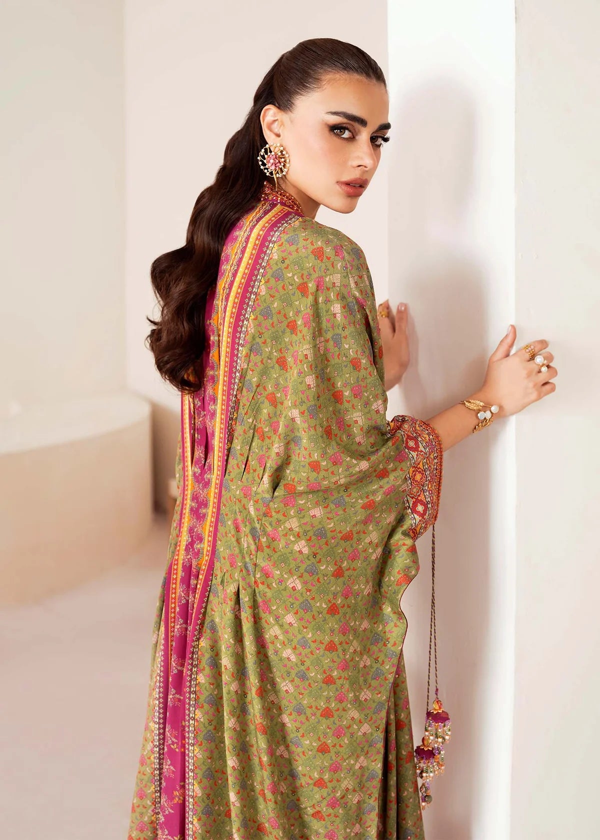 Woman wearing a green floral saree with a pink border, standing against a white wall.