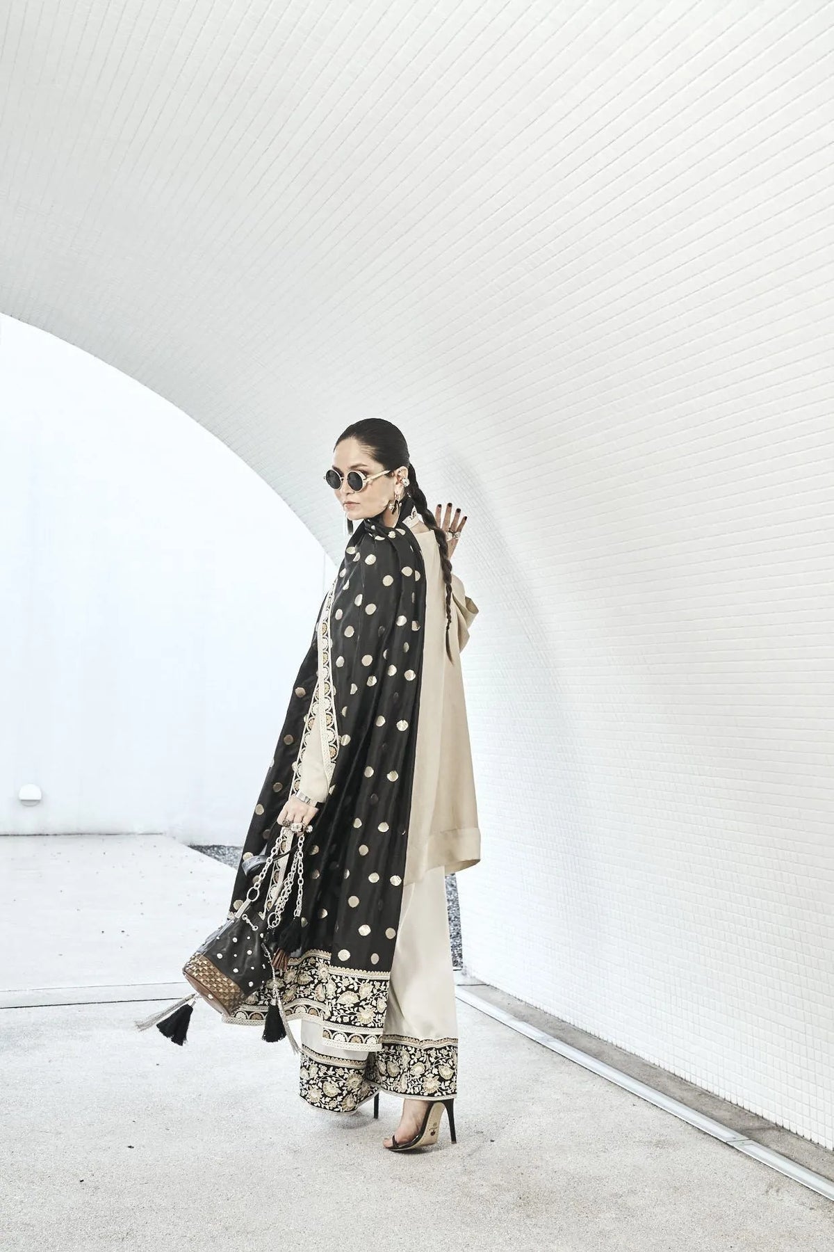 Woman wearing a beige traditional outfit with a black and gold embroidered dupatta, sunglasses, and a matching handbag.