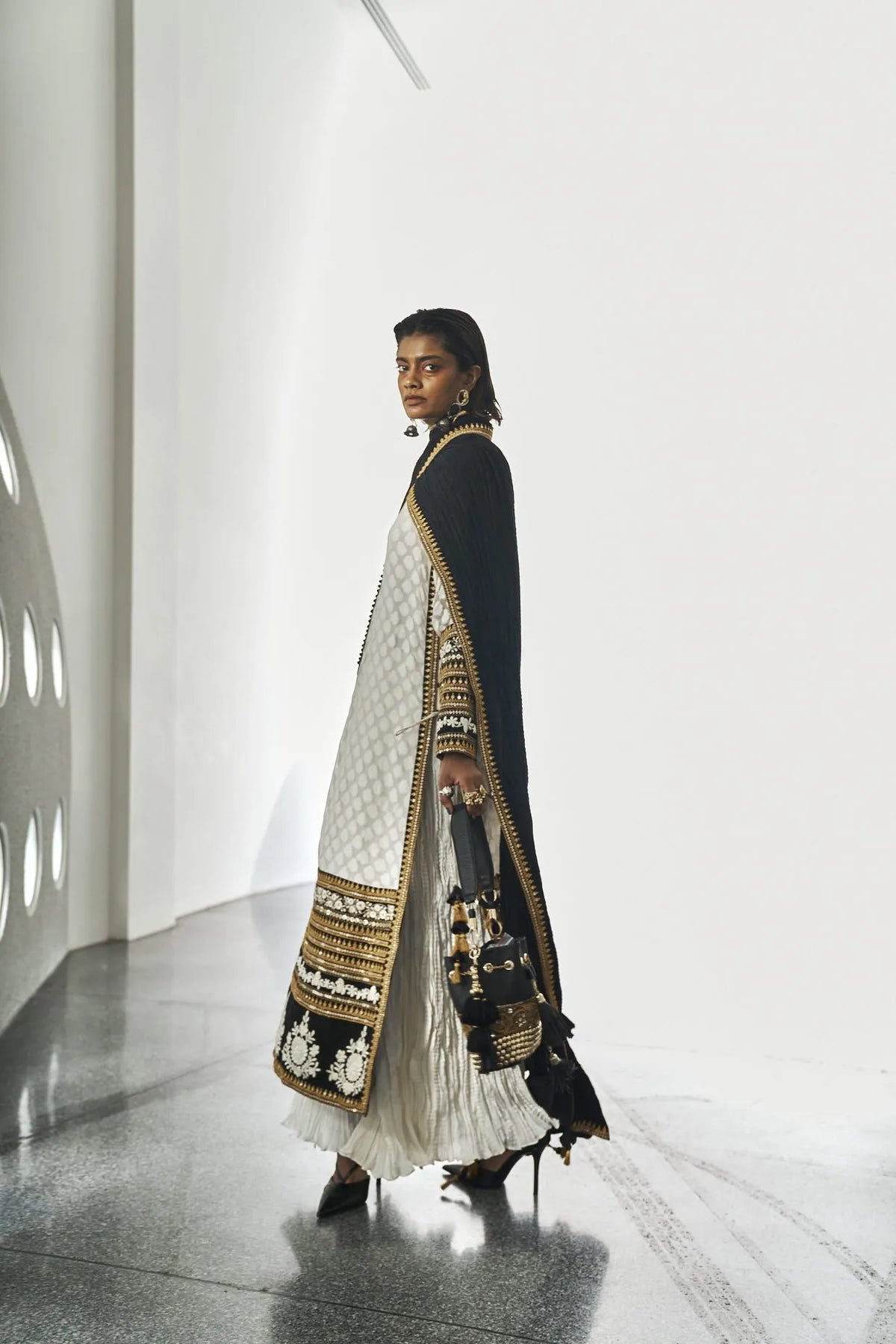 Woman wearing an ivory traditional outfit with black and gold embroidery, a black dupatta, and an embellished handbag.