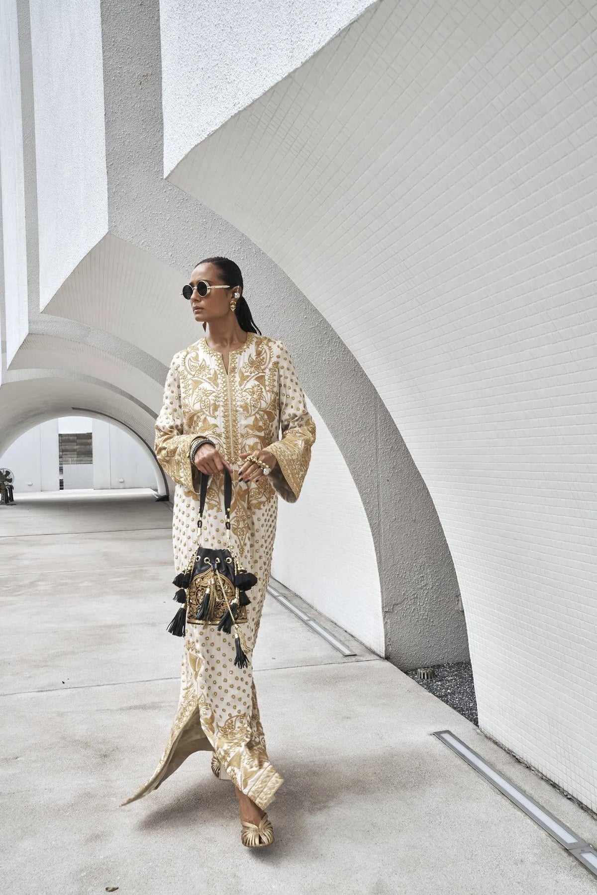 Woman wearing an ivory traditional outfit with ornate gold embroidery, sunglasses, and a black embellished handbag.