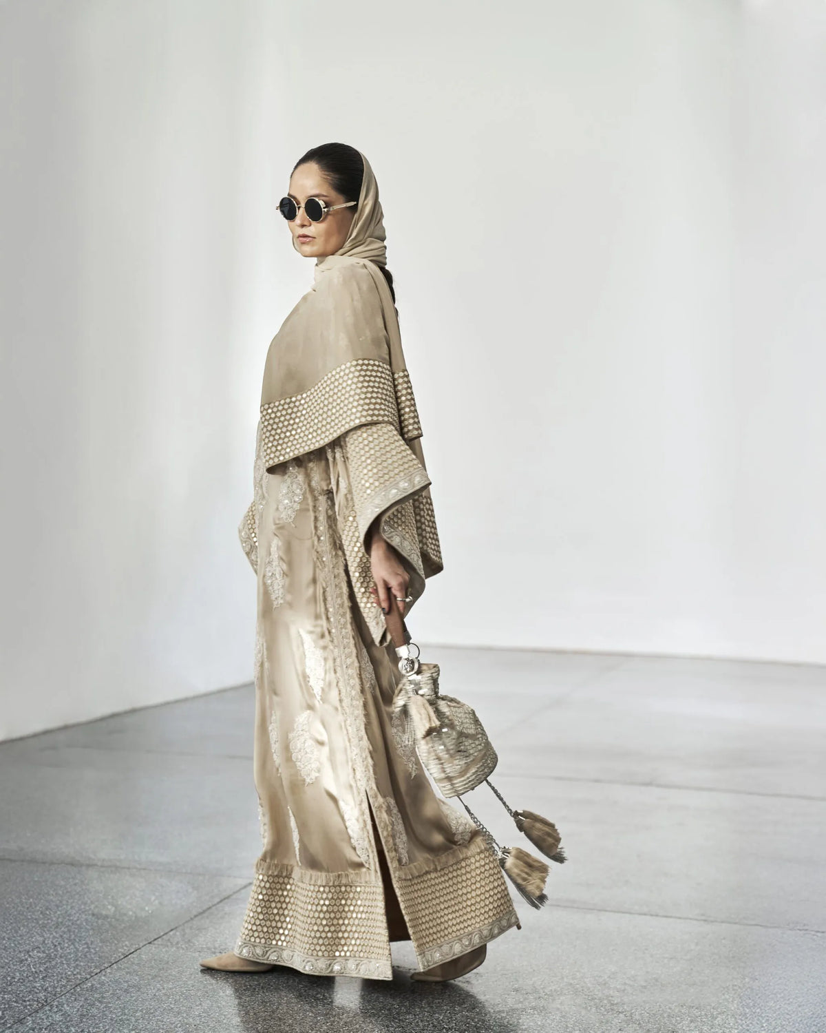 Woman in a beige outfit with a matching headscarf and sunglasses, holding a handbag, against a white background.