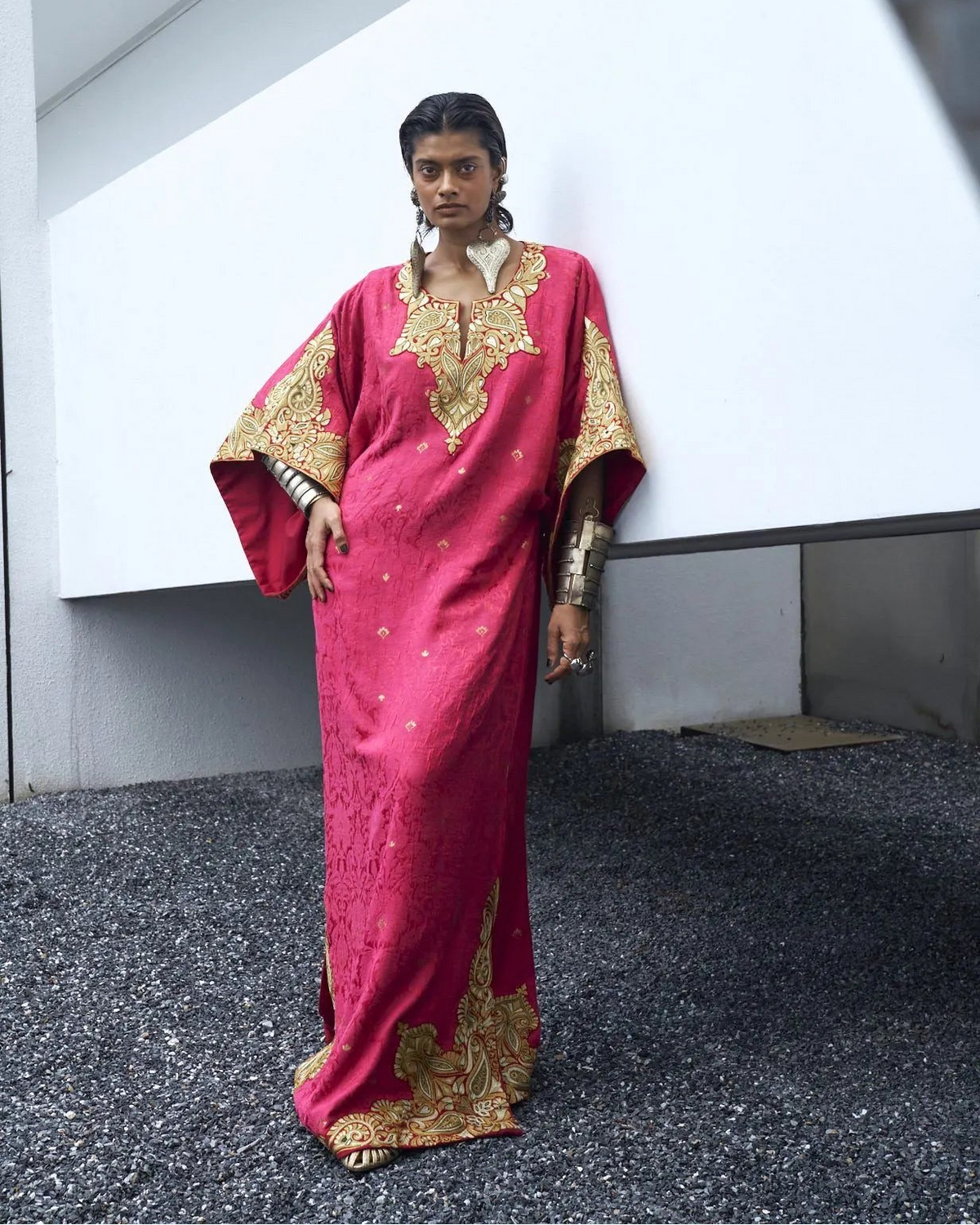 Woman wearing a red and gold traditional outfit standing against a white wall.