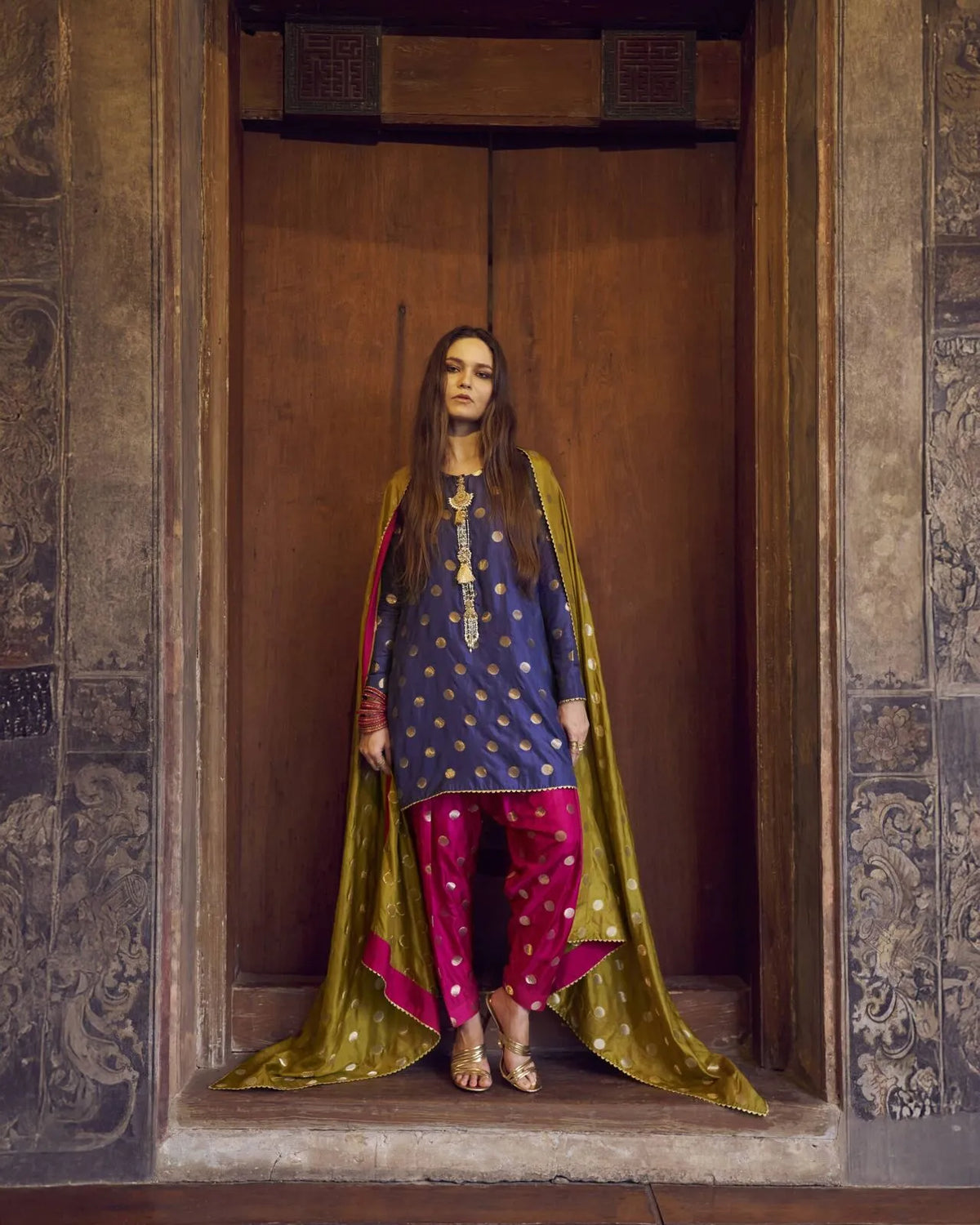 Woman standing in a carved wooden doorway wearing a navy blue traditional kurta with pink trousers and an olive green dupatta.