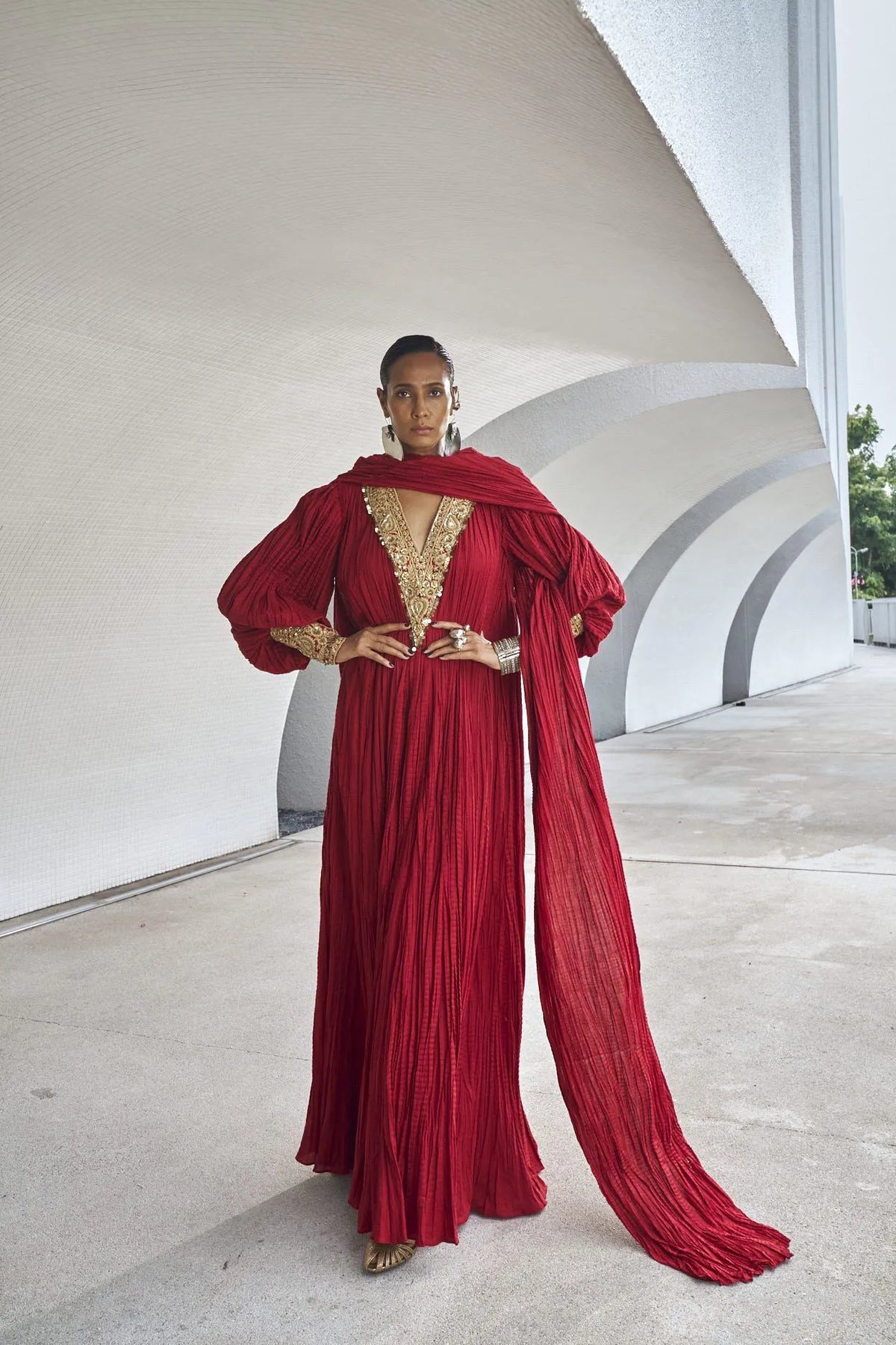Woman standing with hands on her waist in an elegant red pleated dress featuring voluminous sleeves, gold accents, and a draped dupatta.