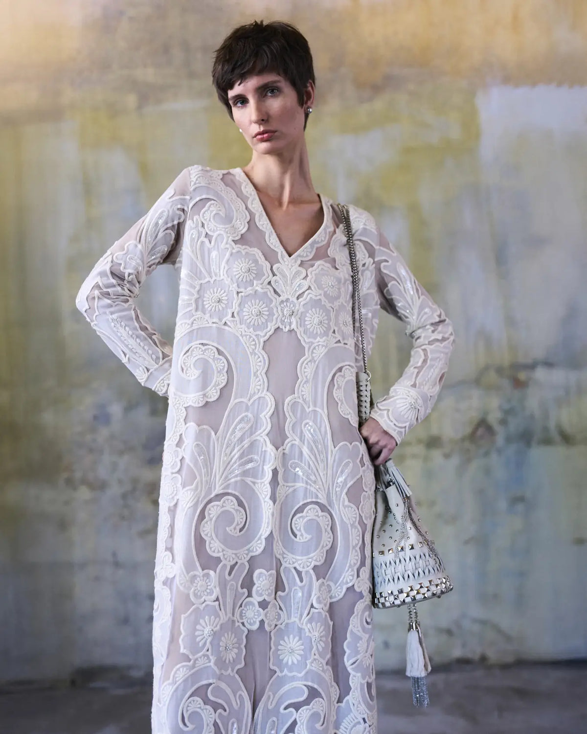 Woman wearing a white lace dress holding a matching handbag against a textured wall.