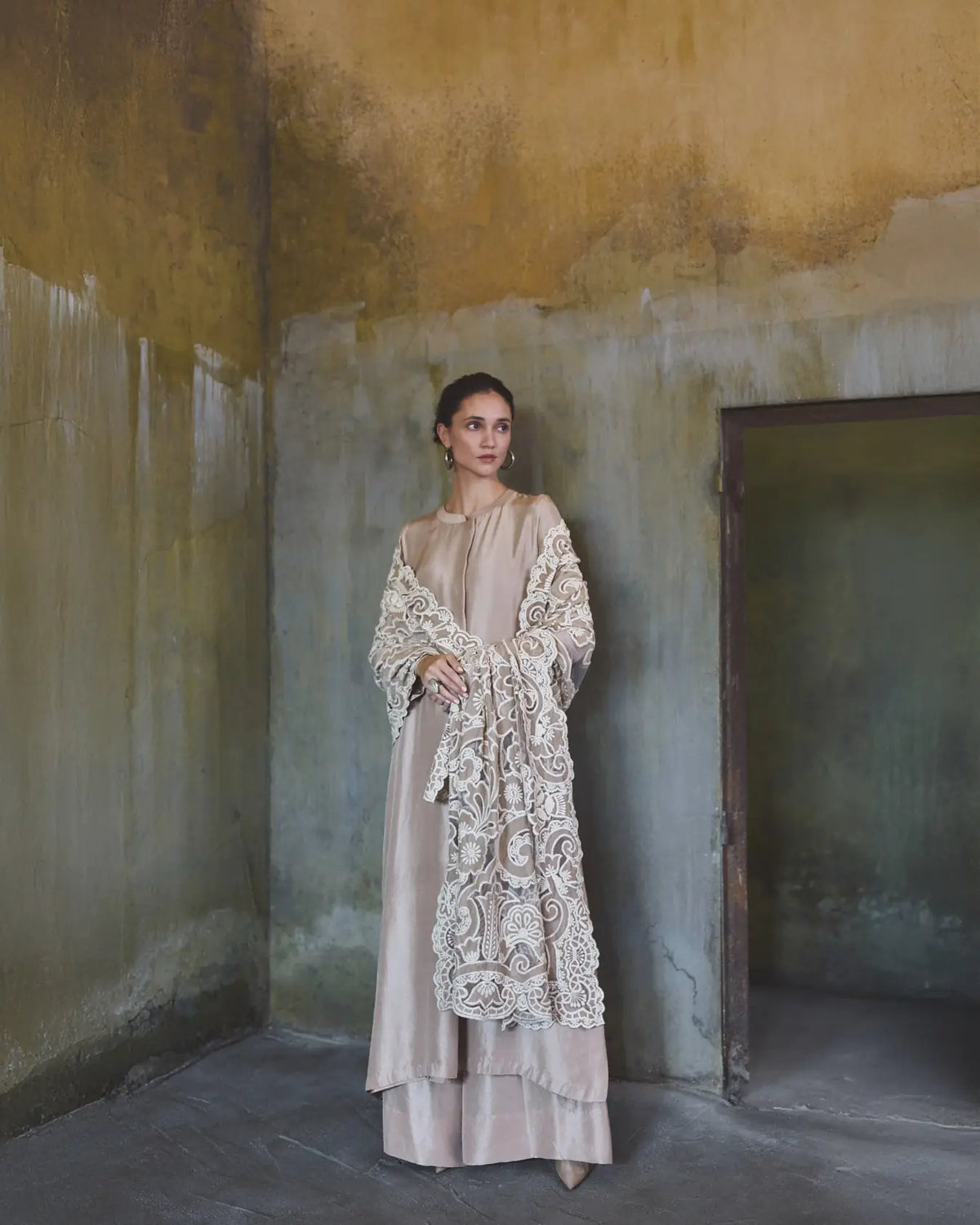 Woman in a beige dress with a long, intricately patterned coat standing in a room with peeling walls.